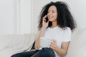 Sideways shot of positive ethnic brunette woman has telephone conversation, drinks hot beverage, holds white cup, sits on comfortable couch in living room, hears pleasant news, smiles happily photo