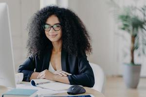 Positive female director with Afro curly hair, rewrites information in notepad from computer, wears optical glasses and formal wear, drinks coffee, poses at desktop, prepares working schedule photo