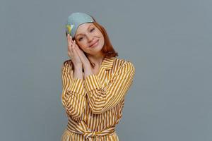 Beautiful relaxed woman tilts head on palms pressed together, looks at camera with satisfaction, enjoys late wake up and having enough sleep, wears pajamas and blindfold, isolated on grey wall photo