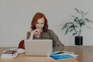 Busy woman freelancer holds spectacles for vision correction, poses at home desktop, has paperwork and works on laptop computer, concentrated at notebook screen. Technology and communication photo