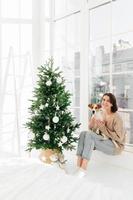 Vertical shot of positive woman poses with jack russell terrier dog, dressed in casual domestic clothes, pose together in bedroom with decorated New Year tree, anticipate for holiday. Winter time