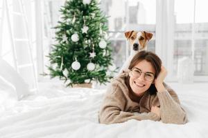 Pretty cheerful woman wears big optical round glasses, lies on bed, her dog poses on back, have fun together, spend winter holidays at home, decorated green Christmas tree. Coziness, winter, festivity photo