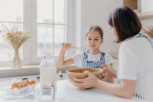 Adorable little child in apron shows whisk with white cream, wears apron, cooks together with mommy, pose in spacious kitchen together, make cake for special event. Children, help about house photo