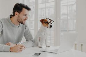 Businessman concentrated in display of laptop computer, writes down notes, works with favourite pet, pose in home office, wear spectacles, work freelance. People, work, technology, animals concept photo