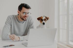 Busy man works freelance, watches training video online on laptop computer wears transparent glasses works together with jack russell terrier pose in home office read news online. Businessman with dog photo