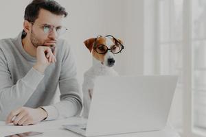 Serious concentrated male freelancer poses in coworking space together with jack russell terrier dog, surf internet, prepare business project together, work from home, wear glasses, read information photo