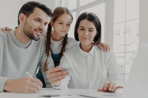 feliz familia amistosa de padre, madre e hija se sientan frente a la computadora, verifican el saldo de la tarjeta de crédito, hacen compras en línea, compran algo necesario, usan conexión a Internet de alta velocidad. foto