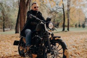 Male motorcyclist drives in nature on fast bike, wears shades, black jacket, gloves, jeans and boots, enjoys autumn season, spends free time actively, ready for long trip. People, transport, driving photo