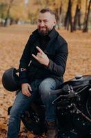 Handsome bearded male motorcyclist holds helmet, makes horn gesture with fingers, feels cool, wears black coat and jeans, sits on fast motorbike against autumn park background. Carefree racer photo