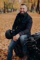 la imagen vertical de un motociclista sonriente sonríe alegremente, sostiene casco, posa en moto, usa chaqueta negra y jeans, conduce por el parque, disfruta de los paseos en otoño en vehículos negros. concepto de transporte foto