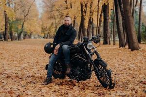 Bearded biker poses on own motorcycle, holds helmet, rides motorbike, poses outdoor in park, ground covered with fallen leaves. Biker lifestyle. Adventure on own transport. Male rider outdoor photo