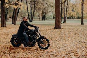 un motociclista seguro de sí mismo se sienta en una moto, disfruta viajar en su propio transporte, usa gafas de sol protectoras, ropa cómoda, pasea por los árboles en el parque de otoño. temporada de otoño, hobby, estilo de vida foto
