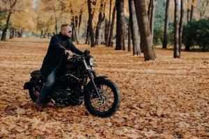 Horizontal outdoor view of active male motorcyclist rides bike, wears trendy sunglasses and black jacket, poses in big autumn park, enjoys awesome travel, has motorcycle trip, rest at nature. photo