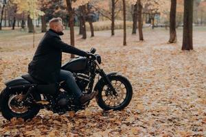 Horizontal shot of bearded biker in action or movement against park during autumn time, going to reach destination quickly, wears sunglasses, jacket, jeans and shoes, has adventurous trip alone photo