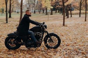 Sideways shot of confident male biker rides motorcycle, has adventure on two wheels in autumn park, wears stylish clothing, protective sunglasses, enjoys nature during beautiful fall season. photo