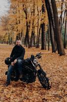 Cheerful biker poses to have break after driving, poses near black motorbike, holds protective helmet, yellow trees and leaves around, enjoys good weather. Autumn season. Urban lifestyle. Outdoor shot photo