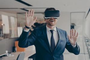 Young excited businessman dressed formally using virtual reality headset while working in office photo