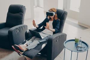 Young modern business woman in VR goggles sitting with opened laptop on armchair at home photo