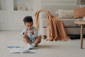 Cute afro american boy kid with colouring book sitting on floor in cozy living room photo