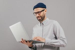 Shot of satisfied male with thick beard and mustache, holds laptop computer, transfers money, uses online banking service, sends files, connected to wireless internet, models over grey background photo