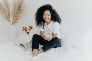 Relaxed attractive Afro American woman drinks aromatic hot drink from white mug, poses on bed together with jack russell terrier dog, enjoy domestic atmosphere, being in cozy bedroom at home photo