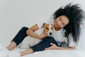 Horizontal shot of lovely curly woman embraces favourite dog, wears whihte t shirt, jeans and socks, enjoy time together, sit on bed. Good beginning of day. Cheerfull lady plays with pet in bedroom photo