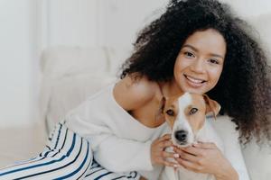 Image of attractive dark skinned woman wears makeup, has bushy curly hair, smiles pleasantly, cuddles dog, dressed in fashionable clothes, enjoys sweet moment with puppy, being at home together photo