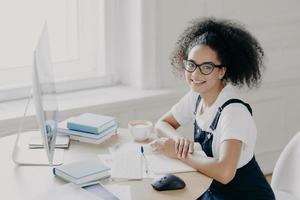 Positive Afro American female freelancer poses at workplace with papers and textbooks, works remotely on computer, has coffee break, works in her own cabinet, wears casual clothes. People, job photo