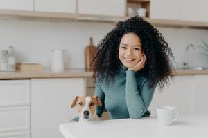 personas, animales, concepto de amistad. una alegre mujer rizada se sienta en la cocina, bebe bebidas calientes, su leal mascota doméstica posa cerca de disfrutar pasando tiempo juntos. chica afro con perro en apartamento moderno foto