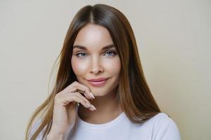 Charming good looking millennial woman keeps hand near face, has healthy glowing skin, dark hair, blue eyes, dressed in casual wear, isolated over brown background. Human face expressions concept photo