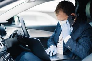 Busy businessman drives car, has phone call and checks information in laptop computer, wears disposable medical mask and gloves to avoid infection of virus. Health care, coronavirus, influenza photo