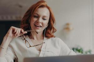 Confident pleased redhead woman concentrated in laptop computer, watches video or movie on laptop computer, enjoys friendly video call, holds transparent glasses, uses free internet connection photo