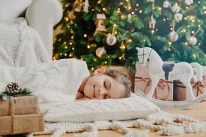 Restful small child lies on soft white pillow, sees pleasant dreams, fall asleep near decorated New Year tree, waits for presents before Christmas Eve, has tender smile on face. Children, coziness photo