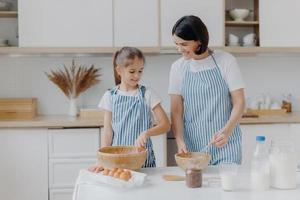 la vista interior de la feliz madre y la hija preparan una cena sabrosa juntas, se paran una al lado de la otra en delantales cerca de la mesa de la cocina, sonríen y baten los huevos en un tazón. gente, familia y concepto culinario foto