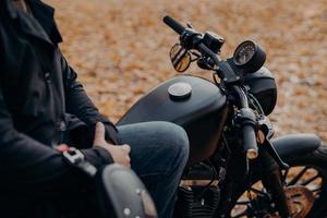 Cropped shot of faceless biker poses on fast motorbike, stops in park, ground covered with fallen leaves, enjoys travel and journey. People, lifestyle, transport concept. photo