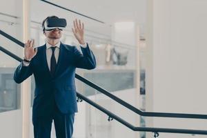 Impressed male office worker wearing VR goggles exploring virtual world while standing in office photo