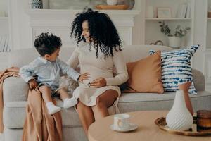 un niño pequeño y lindo tocando el vientre de su madre embarazada mientras se relajaba juntos en el sofá de la sala de estar foto