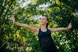 Training outdoors concept. Pleased sporty European woman feels freedom motivates you for sport stretches arms sideways leads active lifestyle wears black t shirt poses against green trees outside. photo