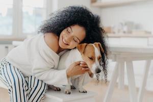 Lovely young curly African American woman embraces beloved pedigree dog with love, has gentle smile, wears stylish clothing, poses against home background in modern apartment, expresses care photo