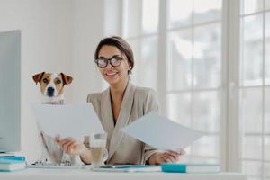 Positive female entrepreneur poses at workplace, holds paper documents, sits in front of computer, wears optical glasses for vision correction, jack russel terrier dog near. Building online business photo