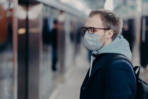 Health care and illness concept. Profile shot of European man wears medical mask, poses on station in underground, takes preventive measures during quarantine, afraids of catching serious virus photo