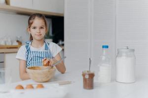 Adorable European child enjoys cooking activity, breaks egg in bowl, whisks ingredients, prepares tasty pastry, being young cook, wears apron, poses against kitchen interior, learns how to make cake photo