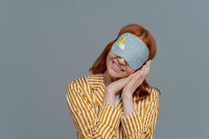 Just relaxing or time for nap. Smiling gentle woman with eye mask, tilts head on pressed palms, wears striped pajama, poses over grey wall, copy space area for your text feels comfortable and relaxed photo