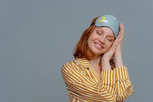 Satisfied ginger woman has nap, sees pleasant dreams, tilts head on palms, smiles gently with closed eyes, wears sleep mask, dressed in striped pajama, isolated over grey background, empty space photo
