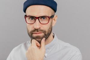 Close up shot of handsome bearded young man holds chin, looks seriously directly at camera, thinks about something, wears spectacles, white clothes, isolated over grey background. Indoor shot photo