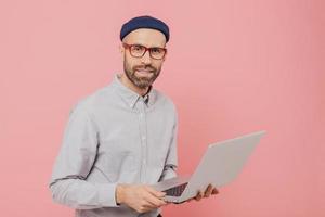 Talented male journalist with appealing look, holds modern laptop computer, works on new article, dressed in fashionable clothes, models against pink background, connected to wireless internet photo