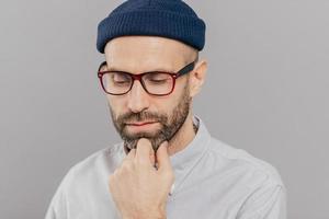 Headshot of sad lonely serious male keeps hand under chin, has dark bristle, looks down, thinks about promotion and starting new career, wears spectacles, hat and shirt, isolated over grey studio wall photo