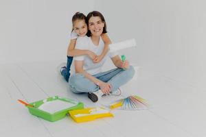 Indoor shot of happy woman gets warm hug from lovely daughter, pose together in empty room, choose bright color for painting from sample palette, busy with house renovation ande redecoration. photo