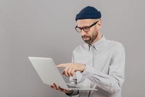 Professional male freelancer makes project work, points with index finger at screen of laptop computer, watches training webinar, holds modern gadget on hands, stands against grey studio wall photo