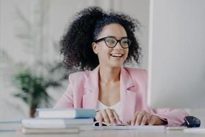 Photo of glad curly professional female manager focused into computer screen, smiles broadly, has curly hair, wears transparent glasses, elegant suit, poses at desktop in her cabinet, works remotely